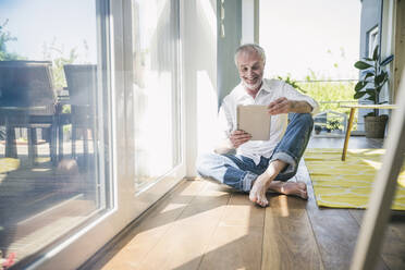 Happy senior man using tablet PC sitting on floor in living room at home - UUF26126