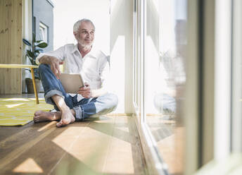 Lächelnder älterer Mann mit Tablet-PC, der zu Hause auf dem Boden sitzt - UUF26129