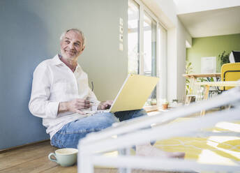 Smiling senior man with laptop and coffee cup sitting on wall at home - UUF26135