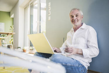 Smiling senior man with laptop leaning on wall at home - UUF26136