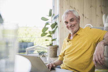 Smiling senior man with laptop sitting in living room at home - UUF26140