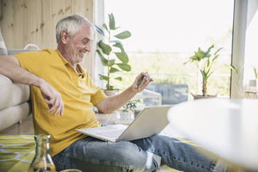 Smiling senior man using smart phone sitting with laptop at home - UUF26141