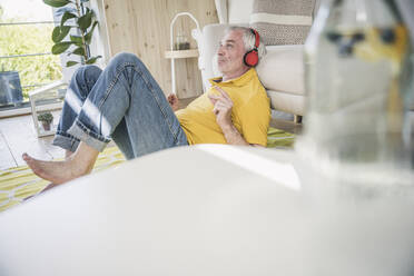 Älterer Mann mit drahtlosen Kopfhörern, der sich zu Hause auf dem Sofa im Wohnzimmer zurücklehnt - UUF26143