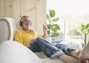 Happy senior man wearing wireless headphones sitting on sofa at home - UUF26144