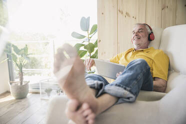 Lächelnder älterer Mann mit drahtlosen Kopfhörern und Tablet-PC auf dem Sofa zu Hause - UUF26146