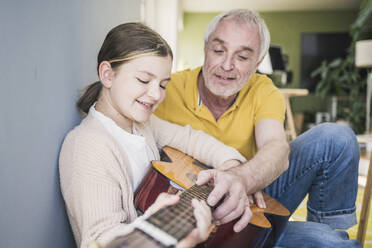 Smiling senior man teaching guitar to granddaughter at home - UUF26149