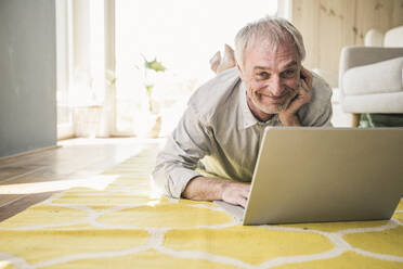 Glücklicher älterer Mann mit Laptop, der zu Hause auf dem Teppich liegt - UUF26155