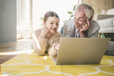 Lächelnder älterer Mann mit Enkelin, die einen Laptop benutzt, der auf dem Teppich im Wohnzimmer zu Hause liegt - UUF26158
