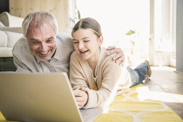Glücklicher älterer Mann mit Enkelin, die einen Laptop auf dem Teppich im Wohnzimmer zu Hause benutzt - UUF26159