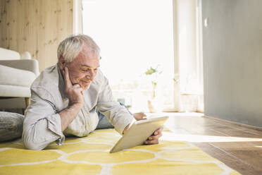 Lächelnder älterer Mann mit Tablet-PC, der zu Hause auf dem Teppich neben dem Sofa liegt - UUF26160