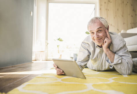 Lächelnder älterer Mann mit Tablet-PC, der zu Hause auf dem Teppich liegt - UUF26161