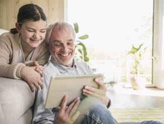 Lächelnder älterer Mann mit Enkelin bei der Nutzung eines Tablet-PCs zu Hause - UUF26163