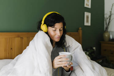 Woman holding tea cup listening music through headphones at home - TYF00152