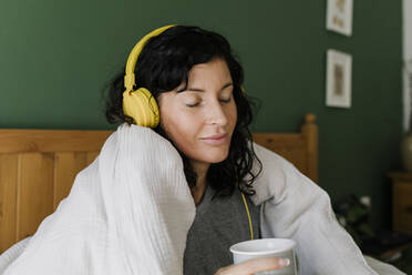 Frau mit geschlossenen Augen, die eine Teetasse hält und über Kopfhörer Musik hört, sitzt zu Hause - TYF00154