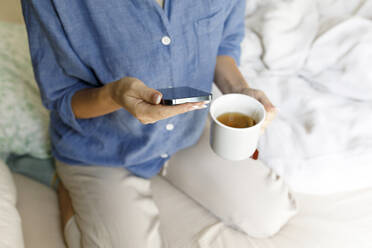 Hände einer Frau mit Handy und Teetasse auf dem Bett sitzend - TYF00162