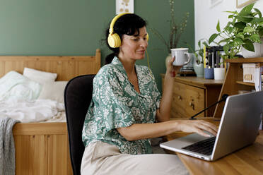 Smiling businesswoman listening music through headphones using laptop at home - TYF00179