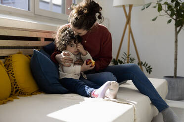 Mother embracing daughter eating apple sitting on sofa - JOSEF09399