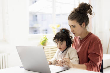 Little girl with headset sitting on lap of mother using laptop - JOSEF09430