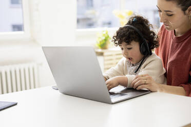 Kleines Mädchen mit Headset sitzt auf dem Schoß der Mutter und benutzt einen Laptop - JOSEF09431