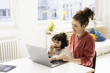 Little girl with headset sitting on lap of mother using laptop - JOSEF09432
