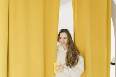 Smiling woman peeking through yellow curtain at home - SEAF00925