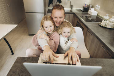Happy businessman with daughters using laptop in kitchen at home - SIF00097