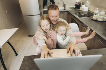 Smiling businessman using laptop sitting with daughters in kitchen at home - SIF00098