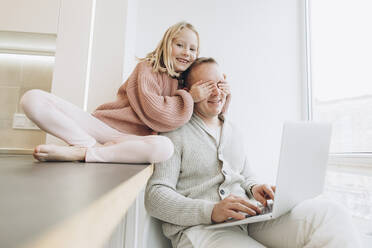 Happy daughter covering eyes of father sitting with laptop by window at home - SIF00119