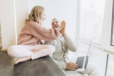 Glückliche Tochter gibt ihrem Vater, der mit seinem Laptop zu Hause am Fenster sitzt, ein High-Five - SIF00121