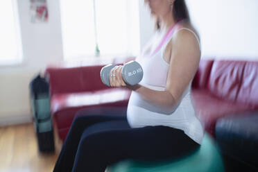 Schwangere Frau, die mit Hanteln auf einem Fitnessball zu Hause trainiert - CAIF32380