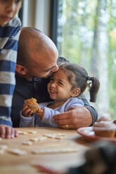 Father hugging happy toddler daughter eating muffin at table - CAIF32414