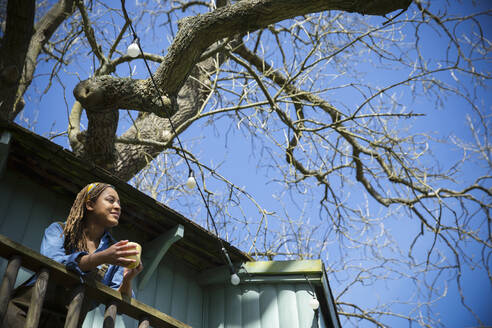 Gelassene junge Frau genießt einen Kaffee auf dem Balkon eines Baumhauses - CAIF32460