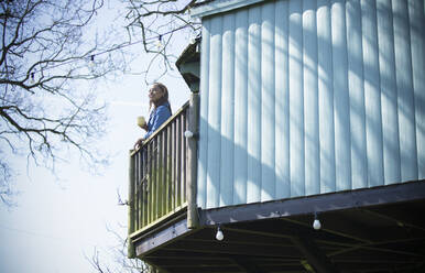 Young woman with coffee on sunny tree house balcony - CAIF32467