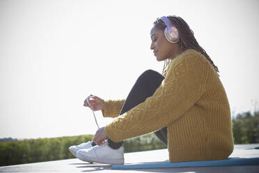 Young woman with headphones tying shoelace outdoors - CAIF32468