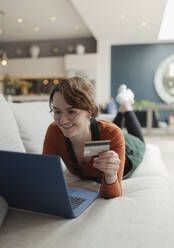 Glückliche junge Frau mit Kreditkarte beim Online-Shopping am Laptop auf dem Sofa - CAIF32494
