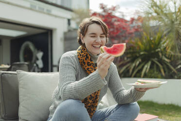 Glückliche junge Frau isst Wassermelone auf einer Sommerterrasse - CAIF32495