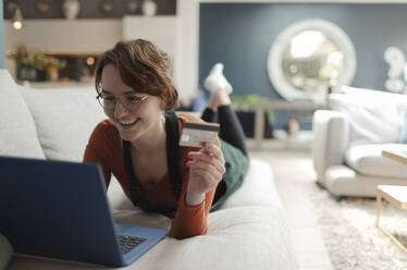 Glückliche junge Frau mit Kreditkarte beim Online-Shopping am Laptop auf dem Sofa - CAIF32497