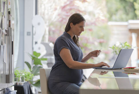 Schwangere Frau mit Kreditkarte beim Online-Shopping am Laptop in der Küche - CAIF32503