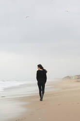 Full body back view of unrecognizable female in black wear strolling on sandy coast near sea during trip in nature - ADSF34715