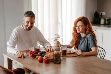 Positive couple having breakfast with strawberries at table with bread in light kitchen at home - ADSF34722