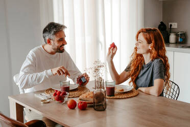 Positive couple having breakfast with strawberries while looking at each other at table with bread in light kitchen at home - ADSF34723