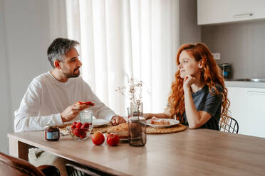 Positive couple having breakfast with strawberries while looking at each other at table with bread in light kitchen at home - ADSF34724