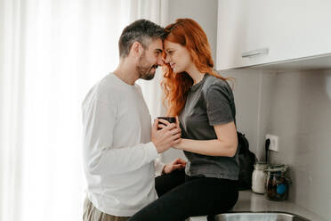 Seitenansicht eines romantischen Paares mit einer Tasse Kaffee, das sich beim Kuscheln auf dem Küchentisch in einer hellen Wohnung ansieht - ADSF34725