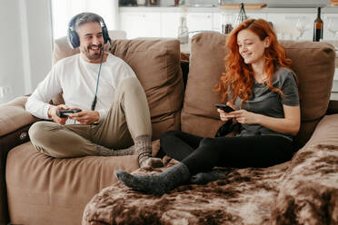 Ganzer Körper eines glücklichen Mannes mit Kopfhörern, der in der Nähe seiner rothaarigen Freundin ein Videospiel spielt, während sie gemeinsam auf dem Sofa im Wohnzimmer sitzen - ADSF34731