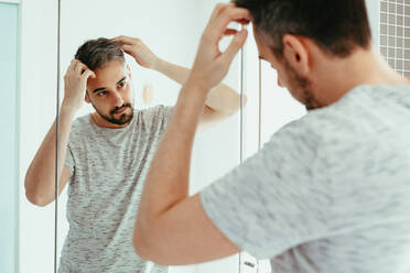Bearded male touching hair while looking at reflection in mirror during daily routine while standing in light bathroom at home - ADSF34735