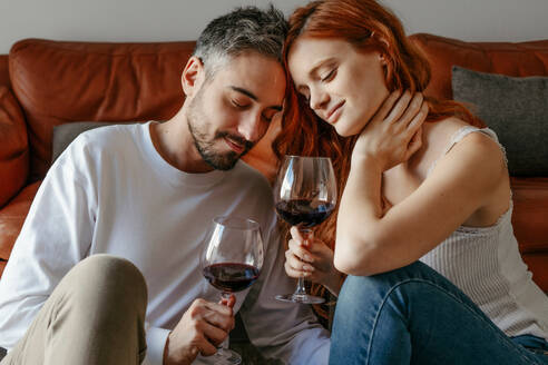 Ganzer Körper eines romantischen Paares mit Weingläsern beim Feiern eines festlichen Ereignisses auf dem Boden in der Nähe der Couch in einem hellen Raum - ADSF34741