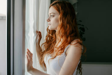 Seitenansicht einer melancholischen jungen Frau mit lockigem rotem Haar, die aus dem Fenster schaut, während sie in einem hellen Raum zu Hause steht - ADSF34744