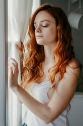 Aufgeregte junge Frau mit lockigem rotem Haar, die in der Nähe von Fenstern in einem hellen Raum mit geschlossenen Augen und unglücklichem Gesicht zu Hause steht - ADSF34745