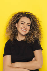Young curly haired woman in black t-shirt looking at camera with arms crossed on yellow background - ADSF34756