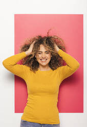 Glückliche junge Frau in leuchtend gelbem Pullover und Jeans, die ihr lockiges Haar auf rosa Hintergrund zerzaust - ADSF34761